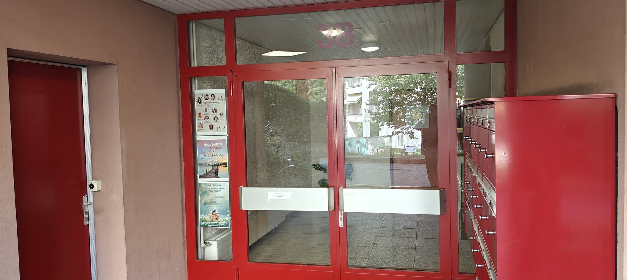 L’entrée de l’immeuble au numéro 38 : une porte vitrée à cadre rouge sous le numéro, les affiches du centre O2 Chemins à gauche